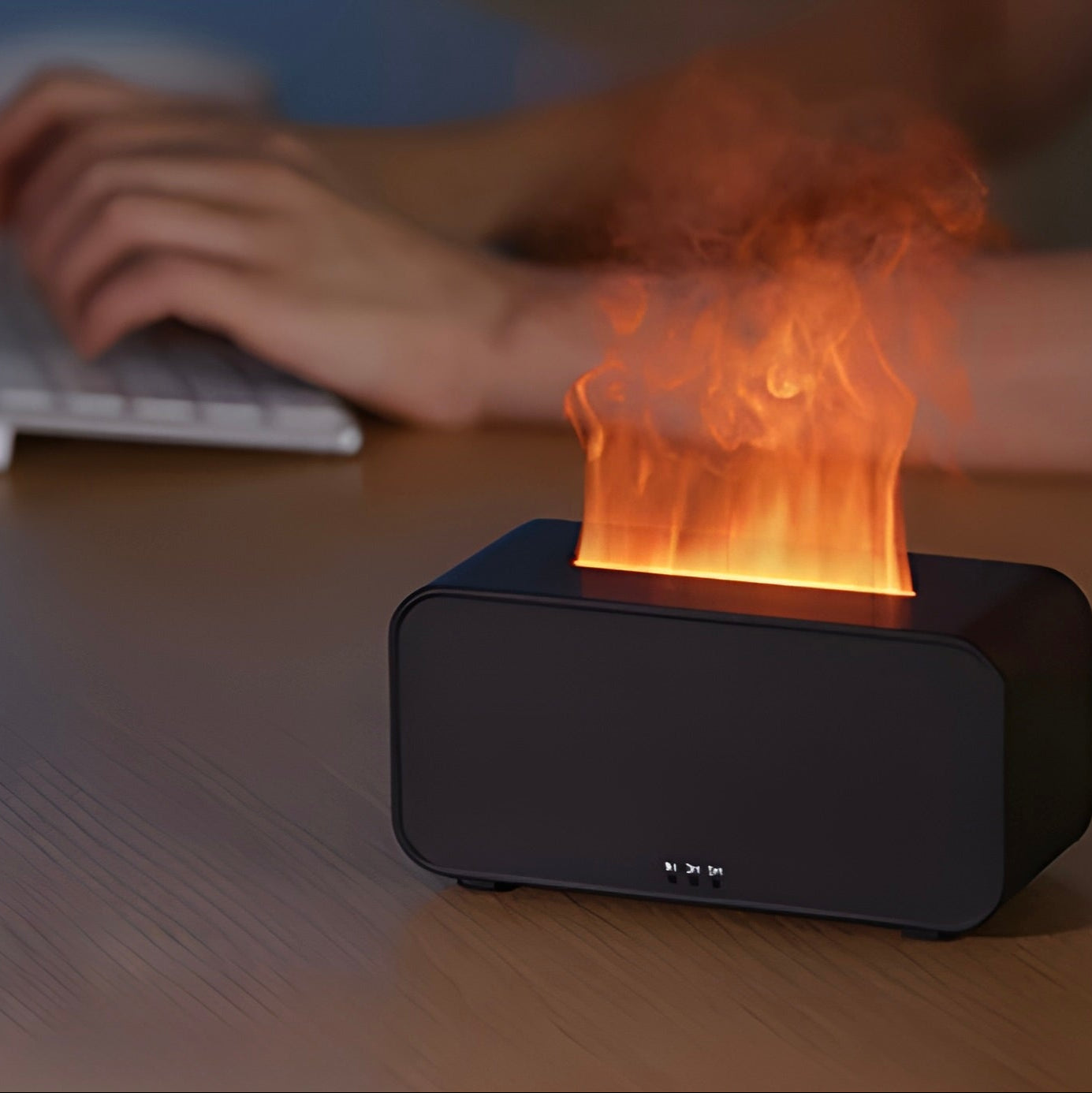Black electronic LED-flame humidifier and diffuser on a wooden desk, person using keyboard in background